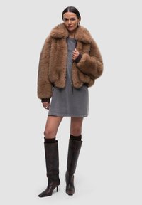 Giacca in pelliccia sintetica marrone con ampio colletto, indossata sopra un vestito grigio. Stivali alti fino al ginocchio, con punta a punta, in marrone lucido con un motivo astratto.