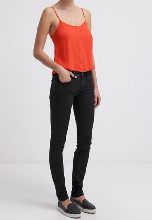 Femme portant un haut sans manches rouge à boutons, un jean skinny noir et des chaussures noires ouvertes, debout avec une main dans la poche.