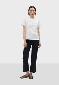 Camiseta blanca de algodón con diseño en relieve 3D que presenta los números "1981". Combinada con pantalones negros y sandalias estampadas. Fondo liso.