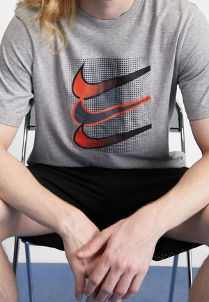 Osoba siedząca, ubrana w szarą koszulkę z trzema nakładającymi się na siebie czerwonymi i czarnymi logo Nike oraz czarne szorty, ręce spoczywają między kolanami.