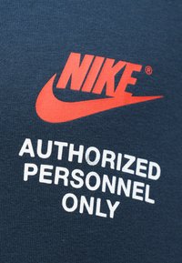 Μπλε υφασμάτινο T-shirt με κόκκινο λογ�ότυπο Nike και το swoosh. Το κείμενο λέει "ΜΟΝΟ ΓΙΑ ΕΞΟΥΣΙΟΔΟΤΗΜΕΝΟ ΠΡΟΣΩΠΙΚΟ" με λευκά γράμματα κάτω από το λογότυπο.
