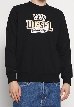 Person trägt einen schwarzen Pullover mit der Aufschrift "1978 DIESEL Industry" in großen Buchstaben, kombiniert mit blauen Jeans.