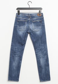 Jeans de mezclilla azul con detalles desgastados y rotos, colgados en un perchero negro sobre un fondo blanco, mostrados desde la parte trasera.