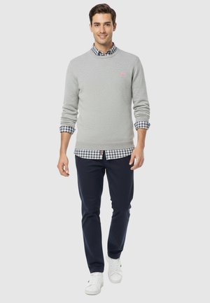 Hombre caminando con suéter gris claro sobre camisa de cuadros en blanco y negro, pantalones azul marino y zapatillas blancas sobre fondo gris.