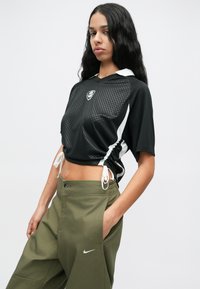 Mulher com cabelo preto longo, vestindo um top desportivo preto curto e calças Nike verde oliva, de pé com uma mão no bolso contra um fundo branco.