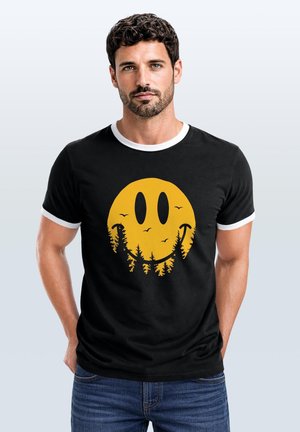 Mann mit dunkel lockigem Haar, der ein schwarzes T-Shirt mit einem gelben Smiley-Gesicht aus Baumsilhouetten und Vögeln trägt, steht mit den Händen hinter dem Rücken.