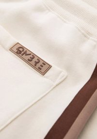 Pantalons en tissu couleur crème avec une ceinture côtelée. Présente une étiquette marron embossée avec "M3RE" et des bandes latérales marron et beige clair.