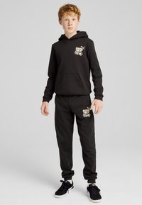 Sudadera negra con bolsillo canguro y logo gráfico en el pecho, combinada con pantalones de chándal a juego que presentan el logo cerca del bolsillo, usados con zapatillas negras.
