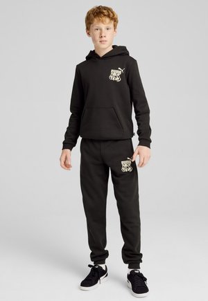 Sudadera negra con bolsillo canguro y logo gráfico en el pecho, combinada con pantalones de chándal a juego que presentan el logo cerca del bolsillo, usados con zapatillas negras.