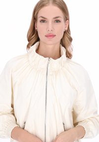 Jachetă de culoarea crem, cu guler adunat, fermoar frontal și manșete elastice. Prezintă o textură fină și un design mulat. Strălucire subtilă.