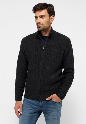Schwarzer Reißverschluss-Pullover mit Fischgrätmuster, gerippten Bündchen und hohem Kragen, über einem marineblauen Hemd getragen und mit blauen Jeans kombiniert.
