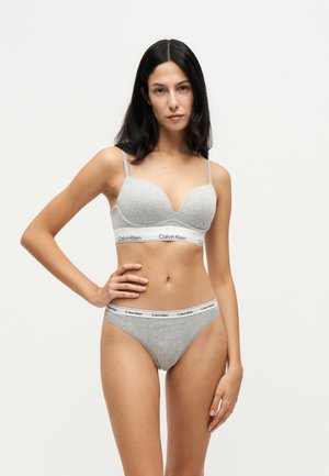 Femme aux longs cheveux bruns portant un ensemble soutien-gorge et culotte gris assorti Calvin Klein, debout, une main sur la hanche, sur un fond clair.