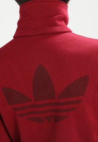 Person som bär en röd polotröja med en stor, mörkröd Adidas trefoil-logotyp på ryggen.