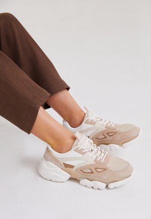Pieds portant des baskets épaisses beige et blanches avec des lacets blancs, associés à un pantalon marron court sur un fond uni.