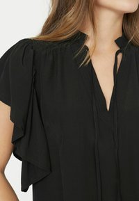 Blusa nera con maniche a farfalla corte, scollatura a V e dettagli di arricciatura sulle spalle. Realizzata in tessuto leggero con una texture morbida.