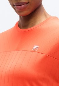 Φωτεινό πορτοκαλί αθλητικό πουκάμισο με υφή ριμπ και στρογγυλή λαιμόκοψη. Διαθέτει μικρό λευκό λογότυπο στο στήθος.