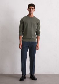 Olive green knit sweater with a round neck, paired with dark blue trousers and black lace-up shoes. Simple design, textured knit material.