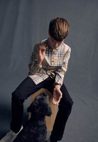 Beiges Karohemd mit Knopfdetails, schwarze Hose und weiße Schuhe. Ein grauer Hund ist in der Nähe der Hand einer auf einem Holzschemel sitzenden Person zu sehen.