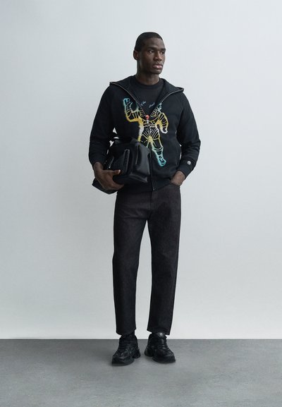 Άνδρας που στέκεται, φορώντας μαύρο φούτερ με στάμπα και σκούρο τζιν, κρατά μαύρη τσάντα, κοιτάζοντας ελαφρώς προς το πλάι σε απλό ανοιχτό γκρι φόντο.