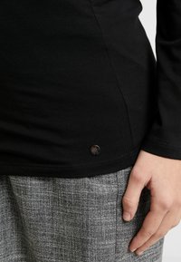 Černá dlouhá rukávová košile s hladkým materiálem a dekorativním černým kovovým knoflíkem na spodním lemu, spárovaná se šedým strukturovaným spodním kouskem.