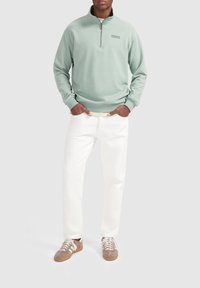 Mintgroene half-zip sweatshirt, zachte textuur, met een klein "BALLIN" logo. Gecombineerd met witte broek en lichtbruine suède sneakers.