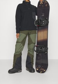 Czarna polarowa bluza z częściowym zapięciem na guziki, zielone i czarne spodnie narciarskie oraz snowboard z czarnymi wiązaniami na neutralnym tle.