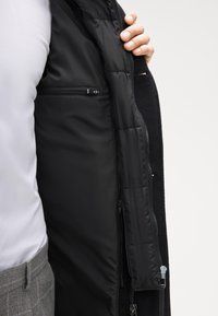 Interno di una giacca imbottita nera con una texture liscia e trapuntata. Presenta una tasca con cerniera e una chiusura con zip robusta. Materiale esterno morbido visibile.