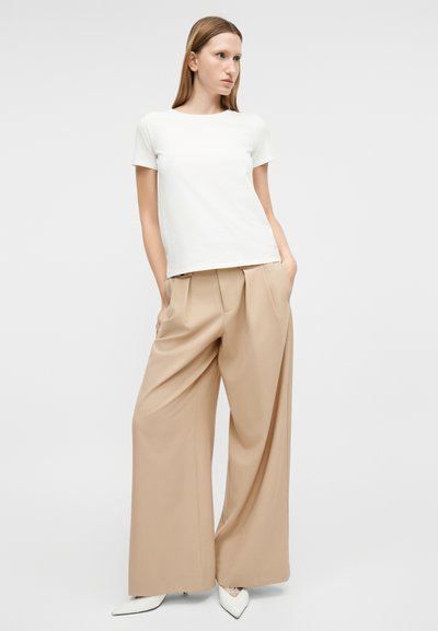 Γυναίκα που φορά λευκή μπλούζα με κοντά μανίκια, μπεζ παντελόνες φαρδιάς γραμμής και λευκά τακούνια με μυτερό άκρο, στέκεται με τα χέρια στις τσέπες κοιτάζοντας πλάγια.