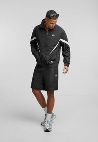 Černá sportovní bunda s bílými detaily, se zipem vpředu a elastickými manžetami, doplněná o odpovídající černé šortky. Model má také na sobě tenisky a ponožky.