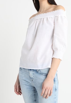 Femme portant un chemisier blanc épaules dénudées avec manches trois-quarts et un jean bleu clair, debout devant un fond uni.