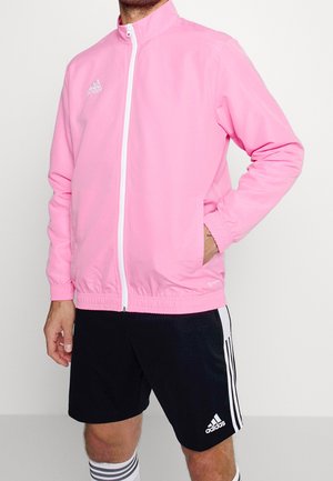 Mies pukeutuneena vaaleanpunaiseen Adidas vetoketjulliseen urheilutakkiin, mustiin Adidas shortseihin valkoisilla raidoilla ja raidallisiin urheilusukkiin, poseeraa yhdellä kädellä taskussa.