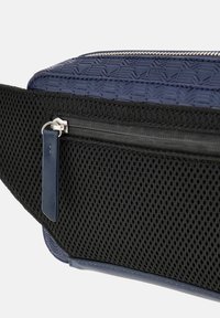 Námornícky modrá ľadvinka s textúrovaným materiálom, sieťovanou vreckom, strieborným zipsom a koženým štítkom, s elegantným a kompaktným dizajnom.