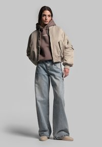Beige bomberjakke over en brun hættetrøje, parret med lys blå vide bukser og beige slip-on sko. Glat struktur, afslappet design.