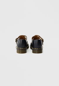 Chaussures Mary Jane en cuir noir avec des brides à boucle et des coutures jaunes, vues de dos sur fond blanc.
