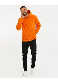 Sudadera de forro polar naranja con un bolsillo tipo canguro y logo en el frente, combinada con pantalones negros de corte ajustado y zapatillas blancas. Textura suave en toda la prenda.