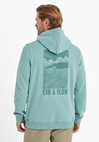 Light teal hoodie with a drawstring, featuring a graphic of clouds and waves in muted tones on the back, with "EBB & FLOW" text.