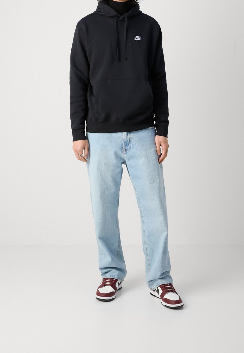 Person som bär en svart Nike-hoodie, ljusblå jeans och vita och vinröda sneakers som står mot en enfärgad vit bakgrund.