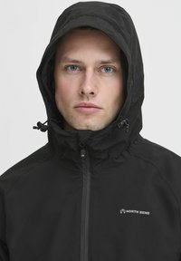 Črna vodoodporna jakna z visokim ovratnikom in nastavljivo kapuco. Ima sprednji zadrgo in vidni logo na levem prsnem delu. Gladka tekstura.