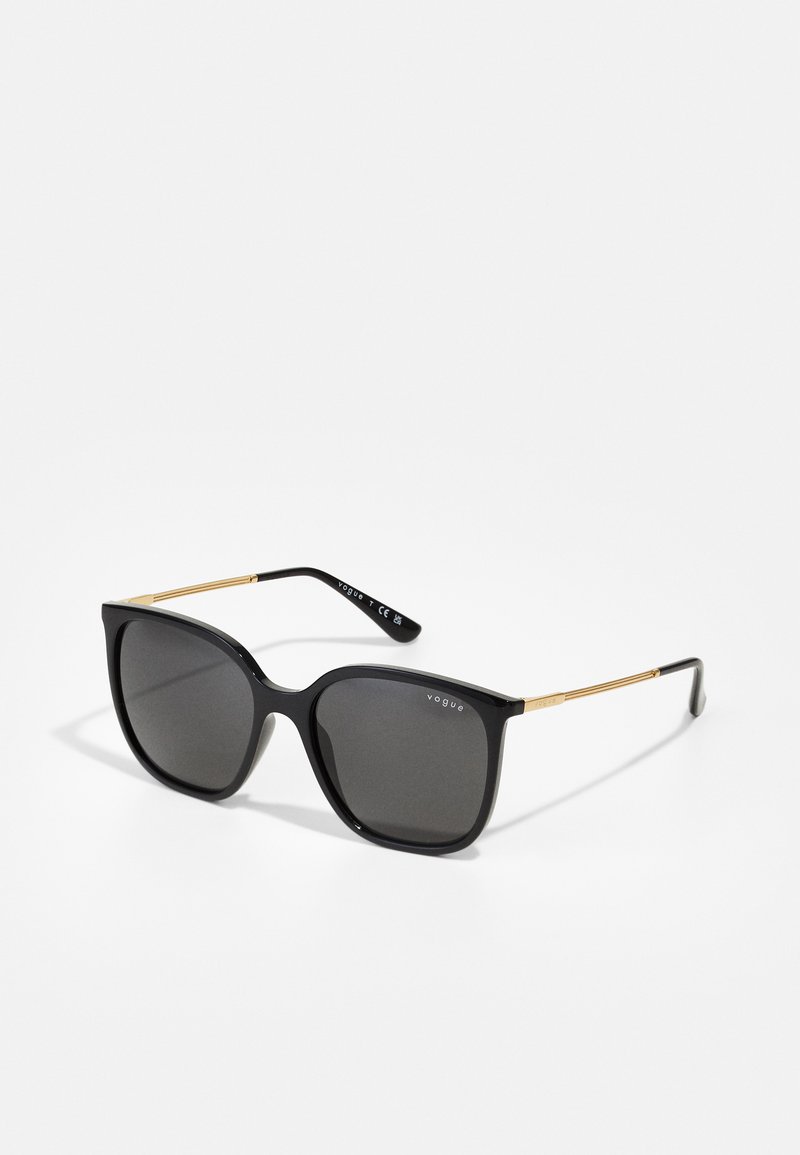 Black sunglasses with a bold, rounded frame, dark lenses, and gold metal accents on the temples. Text reads "vogue" on the upper frame.