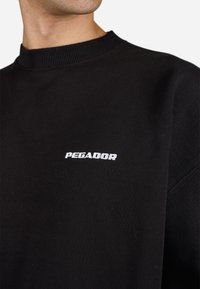 Sudadera negra con cuello redondo y logotipo de texto blanco "PEGADOR" en el pecho, llevada por una persona mostrada desde el cuello hasta la parte superior del torso.