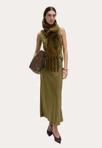 Olivgrön klänning med en texturerad topp; lång scarf med fransar; brun handväska; bruna spetsiga ballerinas; poserar stående mot en neutral bakgrund.