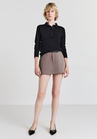Polo noir à manches longues avec patte de boutonnage, associé à une mini-jupe taupe à plis et une fine ceinture noire. Escarpins noirs à bout pointu.