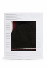 Gorro de lana negro dentro de una caja blanca, con una banda a rayas rojas y blancas en la parte inferior. La caja presenta un diseño minimalista a rayas grises.
