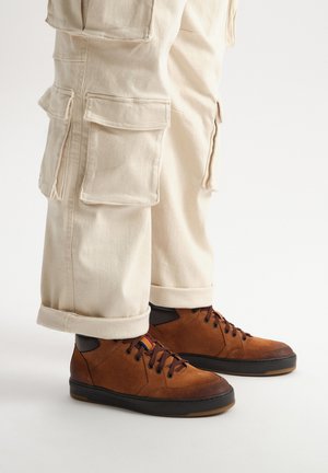 Brązowe zamszowe sneakersy z czarnymi gumowymi podeszwami i sznurowadłami. Wyposażone w wyściełany kołnierz wokół kostki oraz wielokolorowy detal na metce. W połączeniu z beżowymi spodniami cargo.