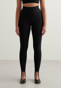 Svarta högmidjade leggings gjorda av ett mjukt, elastiskt tyg med en �åtsittande silhuett och utskärningar vid vristerna, i kombination med svarta skor.