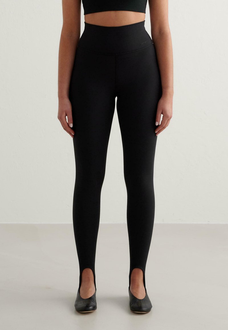 Svarta högmidjade leggings gjorda av ett mjukt, elastiskt tyg med en åtsittande silhuett och utskärningar vid vristerna, i kombination med svarta skor.