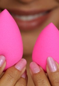Twee roze make-up sponsen vastgehouden in handen, elk met een druppelvorm en een gladde textuur. De achtergrond toont een vervaagde glimlach.