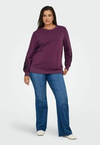 Sweatshirt violet avec broderie florale noire sur les manches, associé à un jean évasé bleu et des baskets blanches. Texture de tissu lisse.