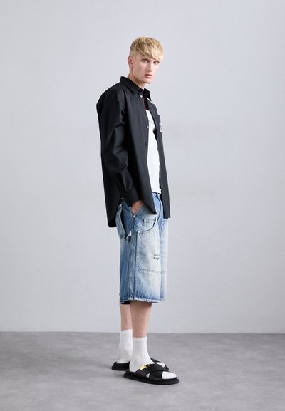 Blond man som bär en svart skjorta över en vit T-shirt, ljusblå denimshorts, vita strumpor och svartasandaler, som står sidledes på en grått golv.