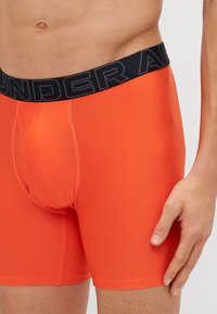 Oransje boxershorts laget av teksturert stoff, med svart elastisk midjebånd med "UNDER ARMOUR"-logo, tettsittende passform og detaljer langs sidesømmen.